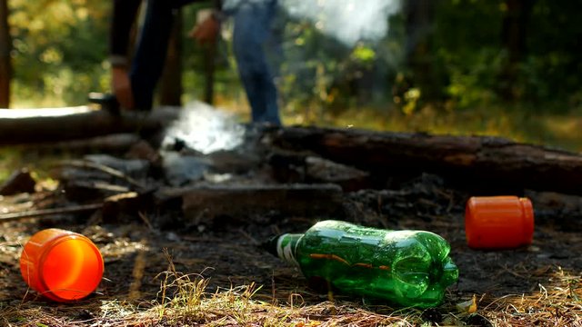 In the foreground in a forest glade there are plastic bottles of garbage, fires are smoking, people walk in the background, outdoor recreation, pollution by garbage