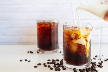 Pouring milk into a glass of homemade cold brew coffee on white table background.
