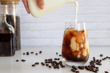 Pouring milk into a glass of homemade cold brew coffee on white table background