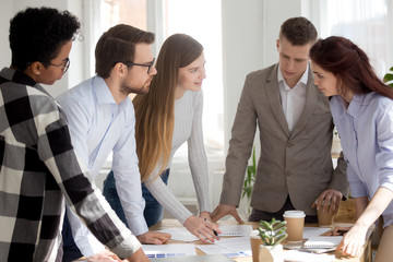 Five confident multiracial millennial diverse business people standing surrounding desk discussing in office boardroom. Young designers solving making a decision, working together at meeting briefing