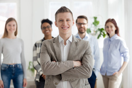 Smiling millennial confident businessman stands with arms crossed looking at camera with happy cheerful business partners team on background. Successful teamwork, company owner and leadership concept