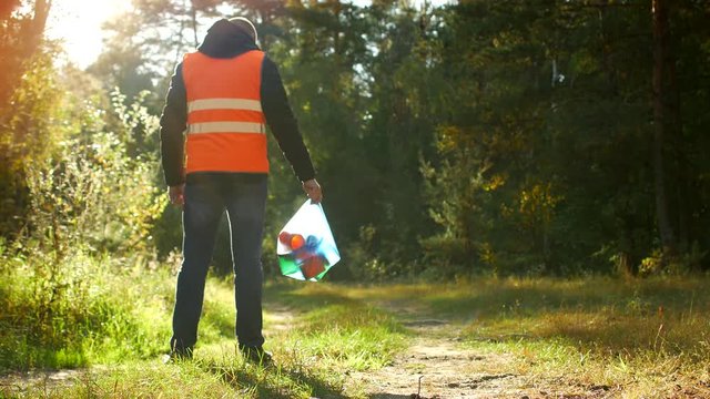 A man in a signal orange vest is standing with a package of garbage in his hands in a forest glade, nature and garbage, garbage collection in the forest, a volunteer, rubbish
