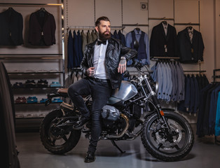 Stylish tattooed bearded man with dressed in black leather jacket and bow tie posing near retro sports motorbike at men's clothing store.