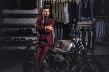 Obraz na płótnie Canvas Handsome man with a stylish beard and hair dressed in vintage red suit posing near retro sports motorbike at men's clothing store.