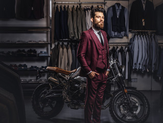 Fototapeta na wymiar Handsome man with a stylish beard and hair dressed in vintage red suit posing near retro sports motorbike at men's clothing store.