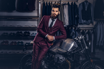 Handsome man with a stylish beard and hair dressed in vintage red suit posing near retro sports motorbike at men's clothing store.