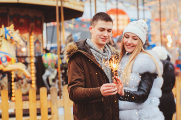 happy couple with christmas firelights walking in city holiday amusement park, carousel on background