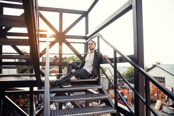Young handsome hipster man with skateboard on the black iron stairs of the industrial building.
