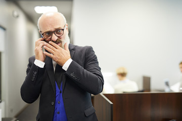 Disgusted or confused senior man in formalwear talking by smartphone in conference hall