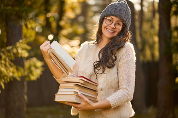 beautiful woman in a cap and glasses is holding a pile of books on the background of autumn forest