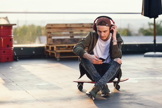 Young handsome man listening music in headphones and using smartphone while sitting on skateboard on the rooftop of industrial building at the sunset. - Powered by Adobe