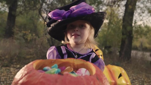 a little beautiful girl in a witch costume and hat takes candy from the bucket. Halloween in the autumn Park.