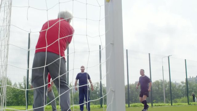Group of four senior men playing with football outdoors on warm summer day, one of them standing at goal net and catching ball, his teammate clapping hands
