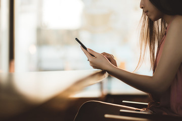 Young asian woman using mobile phone at sunny office while sitting on wooden chair.