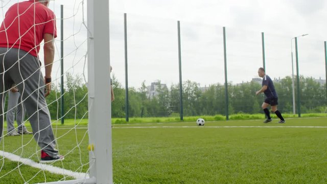 Slow motion shot of group of four active retired men playing with football on warm summer day, one of them kicking ball and one failing to catch it while standing at goal net