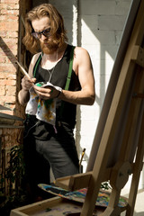 Portrait of contemporary long-haired artist wiping brushes while painting by easel in art studio lit by sunlight , copy space