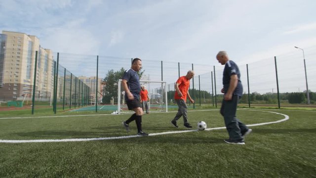 Group of active elderly people in sports clothes playing football outdoors on warm summer day, two teams competing with each other and trying to score goal