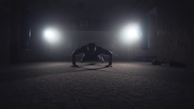 Professional boxer doing push-ups in dark smoky gym. Athlete training in elastic bandages. Sport concept in slow motion. Silhouette on dark background