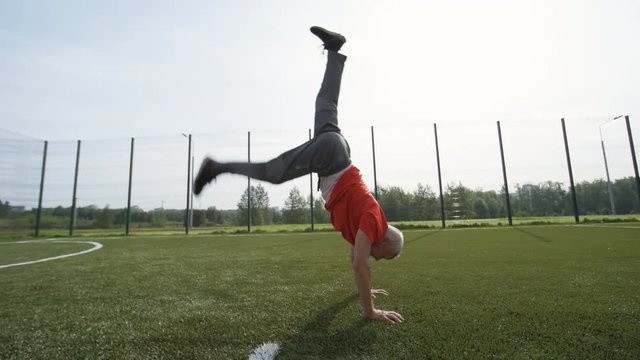 Side view of energetic elderly man exercising on outdoor football field and performing handstand