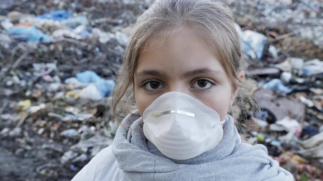 Young sad girl in a white respirator on a dump looks into the camera. Ecology concept.