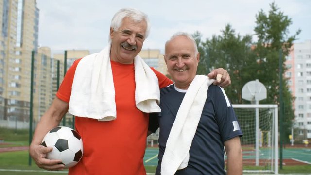 Medium shot of two optimistic elderly male friends with towels on shoulders hugging and talking to each other while standing on football field after game, one of them holding ball