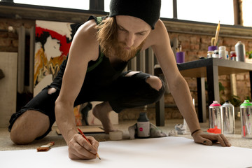 Full length portrait of inspired contemporary male artist sketching picture with pencil on floor in art studio, copy space