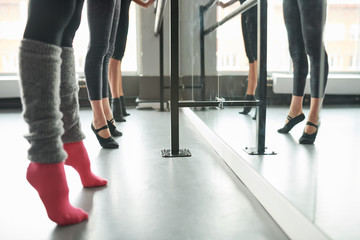 Low section view at row of elegant young women practicing ballet moves standing by bar against mirror in dance studio