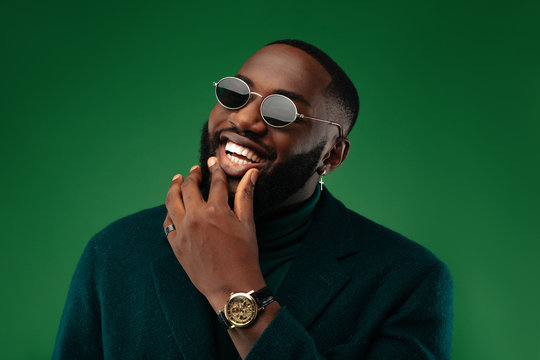 Man portrait. Style. Handsome Afro American guy in green jacket and sun glasses is looking at camera and smiling, on a green background