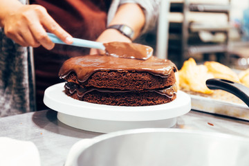 Baker chef preparing moist and spatula dark chocolate torte cake and putting the top with cocoa cream mixed together in kitchen in bakery shop.
