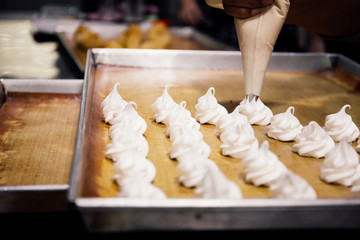 Human using pastry or piping bag to squeeze out white whipped cream of meringues on bakery tray in...
