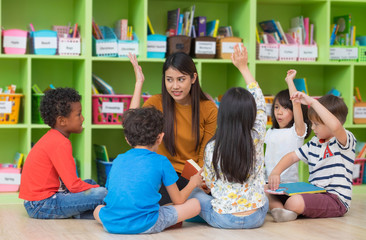 Asian female teacher teaching and asking mixed race kids hand up to answer in...