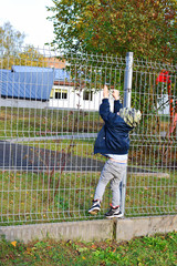 Little three years old boy is climbing the fence while walking on street in autumn day.