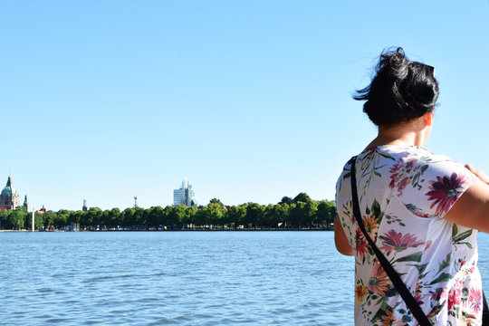 Woman Looking Around On Landscape With Trees And City Across Lake Back View.