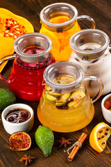 Vertical iimage of glass teapots set with colorful natural hot tea decorated with frruits and jam at wooden dark board background.