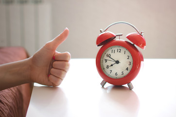 Sleepy woman in the bed is showing a thumbs up gesture by her hand to a red alarm clock on the...