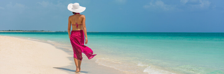 Luxury travel vacation elegant lady walking on beach in pink fashion skirt wrap relaxing on Caribbean holidays during winter. Panoramic banner landscape crop for background. - obrazy, fototapety, plakaty