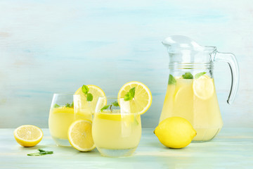 A side view of homemade lemonade in glasses and a pitcher, with fresh lemons and mint, on a teal...