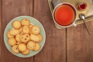 An overhead photo of Danish butter cookies on a plate, on a dark rustic background with a cup of tea on a vintage tray, with jam, and a place for text