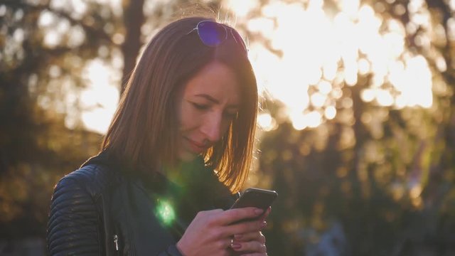 Attractive young girl stands in middle of busy buzzling city on summer sunset. Gorgeous young woman in a bright sunlight uses her phone.