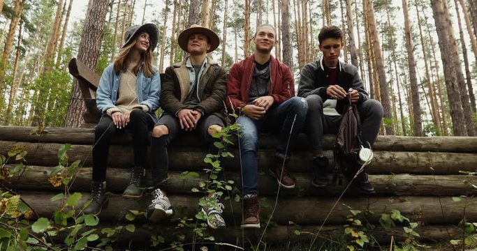 Four young people in jeans and jackets sitting on pile of logs in forest on background of trees. One guy is sad he is offended