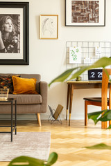 Posters above sofa and desk with laptop in freelancer's interior with wooden chair. Real photo