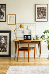 Wooden chair at desk with lamp and laptop in white home office interior with posters. Real photo