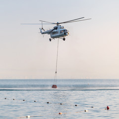 A helicopter of the fire service with a fire fighting bucket is taking part in putting out a fire. A helicopter with a red basket is lowered over the sea to catch water.
