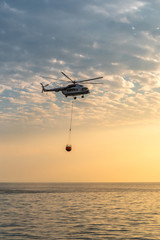A fire helicopter collects water into the sea and flies toward the mountains to extinguish a forest fire on the background of a beautiful dawn sky. Rescue operation. Forest Fire Prevention