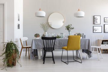 Black and yellow chair at table in white dining room interior with plants, lamps and mirror. Real photo - obrazy, fototapety, plakaty
