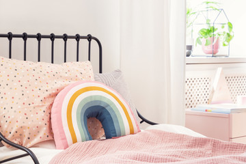 Rainbow pillow and pink sheets on girl's bed in bright bedroom interior with plant. Real photo - obrazy, fototapety, plakaty