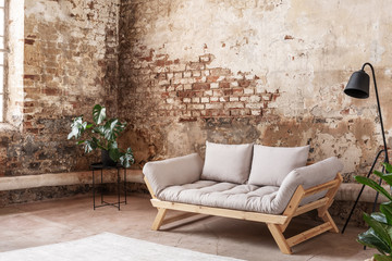 Grey sofa between plant and black lamp in wabi sabi loft interior with red brick wall. Real photo