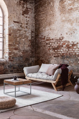 Pouf and wooden table on carpet in industrial loft interior with grey sofa against red brick wall....
