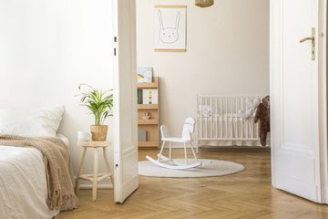 Plant on stool next to bed in white bedroom interior with rocking horse on rug and cradle. Real...