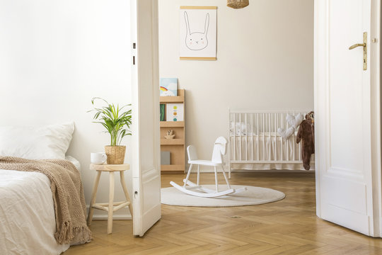 Plant on stool next to bed in white bedroom interior with rocking horse on rug and cradle. Real photo